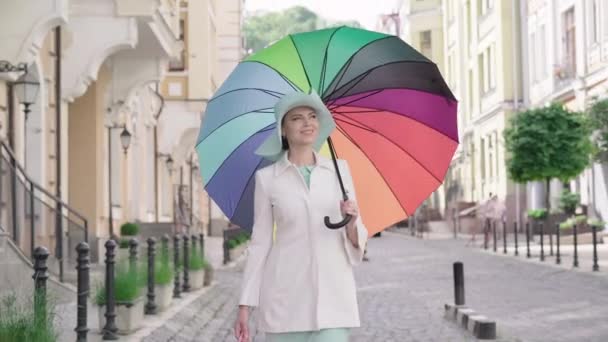 Bella donna in elegante cappotto e cappello passeggiando lungo la strada della città europea con ombrellone colorato e borse della spesa. Ritratto di donna felice che sorride e chiude l'ombrello. Moda, stile. — Video Stock