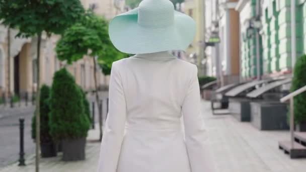 Kamera podąża za pewną siebie modną damą spacerującą po słonecznej miejskiej ulicy, zwracając się do kamery i uśmiechając się. Elegancka, stylowa biała kobieta spacerująca z torbami na zakupy w europejskim mieście. — Wideo stockowe