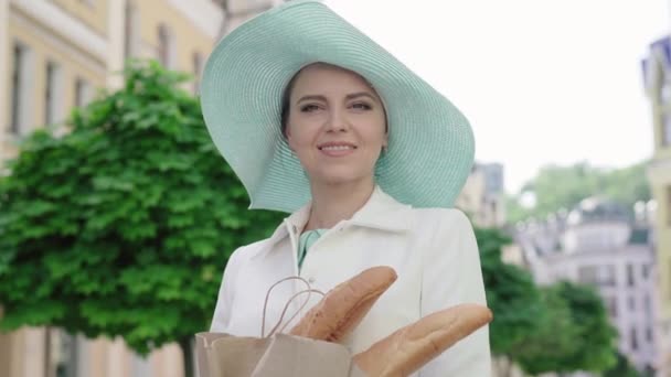 Portret van een charmante vrouw met een elegante hoed die op straat staat met stokbrood en rondkijkt. Prachtige blanke vrouwelijke toerist die geniet van een zonnige dag in de Europese stad. Toerisme, vrije tijd, vreugde. — Stockvideo