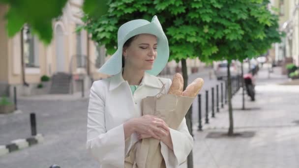 中间一张优雅的女人撕破一块面包，对着相机微笑的照片。迷人的中年白人女士在阳光灿烂的欧洲城市里享受旅行的画像。快乐、旅游、生活方式、优雅. — 图库视频影像