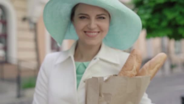 Wazige blanke vrouw die verse, smakelijke stokbrood uitrekt tot camera en glimlacht. Portret van een elegante positieve dame poseren met bakkerij op zonnige stad straat buiten. — Stockvideo