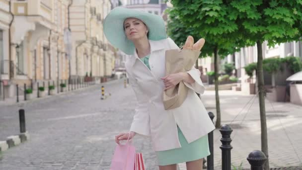 Elegante elegante mujer caucásica autoestop en la calle de la ciudad europea. Retrato de dama adulta segura parada al aire libre con bolsas de compras y baguettes. Estilo de vida, turismo, ocio . — Vídeos de Stock