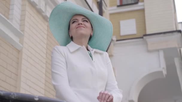 Portré mosolygós pozitív kaukázusi hölgy elegáns kabát és kalap áll a szabadban a városban, és néz félre. Középső lövés elegáns bájos nő élvezi napsütéses nap. Turizmus, utazás, szabadidő. — Stock videók