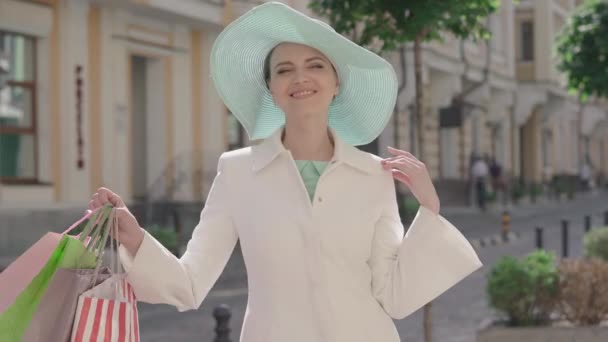 Morena de olhos castanhos mulher esticando sacos de compras para câmera e sorrindo. Retrato de uma senhora caucasiana adulta a gabar-se de compras na ensolarada rua da cidade. Moda, alegria, estilo de vida . — Vídeo de Stock