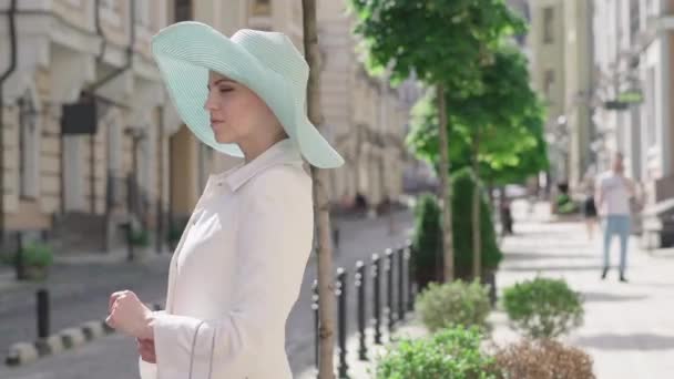 Vista laterale di fiduciosa donna di mezza età guardando bottiglia di vino bianco sulla strada della città e lasciando. Ritratto di elegante signora caucasica in cappello e cappotto che va in albergo con gli acquisti. Turismo, tempo libero, gioia. — Video Stock