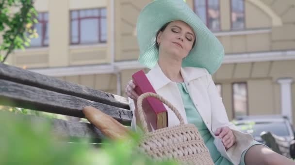 Portrait de femme confiante assise sur un banc en ville et livre d'ouverture à lire. Élégante dame caucasienne appréciant la lecture en plein air le jour d'été ensoleillé. Joie, passe temps, style de vie. — Video
