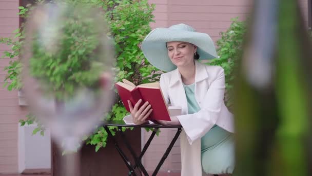 Stilvolle kaukasische Frau liest Buch und lächelt, als säße sie im Café. Vorne stehen verschwommene Flasche und Weinglas. Porträt einer eleganten Touristin, die den sonnigen Tag im Freien genießt. — Stockvideo