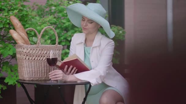 Femme souriante dans un élégant chapeau turquoise lisant le livre et buvant du vin rouge à l'extérieur. Portrait de dame blanche confiante reposant avec de l'alcool et de la littérature sur une journée ensoleillée en ville. — Video