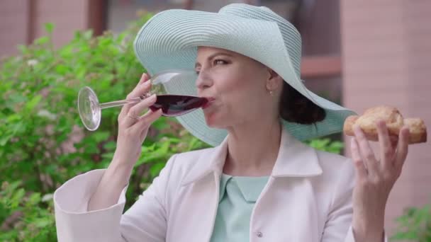 Portret van een aantrekkelijke vrouw die rode wijn drinkt en buiten lekker croissant ruikt. Positieve Kaukasische dame met elegante hoed genieten van vakanties in de Europese stad. Toerisme, vrije tijd, vreugde, levensstijl. — Stockvideo