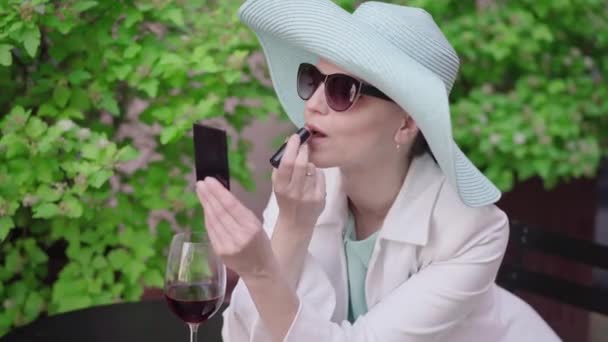 Портрет стильной женщины в бирюзовой шляпе и солнцезащитных очках с помадой. Красивая кавказская леди, любующаяся отражением в ручном зеркале, сидящая на улице с бокалом красного вина. Досуг, красота . — стоковое видео