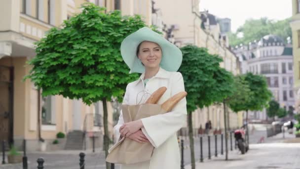 戴着雅致帽子的时髦女士站在城市街道上,手里拿着面包.在阳光明媚的夏日，一个长相像法国人的中年白人女人在欧洲旅行。快乐、生活方式、旅游、快乐. — 图库视频影像
