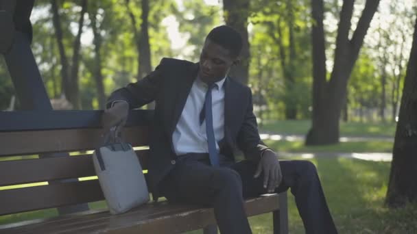 Hombre afroamericano cansado en traje de negocios poniendo la bolsa en el banco y mirando hacia otro lado. Retrato de un joven empresario exitoso teniendo un descanso en el soleado parque de verano. Ocio, riqueza, estilo de vida . — Vídeos de Stock