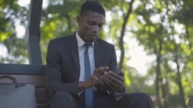 Yaz parkında bankta oturan akıllı telefonu kullanan genç bir adam. Kendine güvenen Afro-Amerikan iş adamının portresi internette sörf yapıyor ve gülümsüyor..