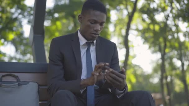 Yaz parkında bankta oturan akıllı telefonu kullanan genç bir adam. Kendine güvenen Afro-Amerikan iş adamının portresi internette sörf yapıyor ve gülümsüyor.. — Stok video
