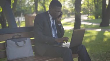 Meşgul Afrikalı Amerikalı işadamı telefonda konuşuyor ve dizüstü bilgisayarında klavye çalıyor. Yaz parkında online çalışan başarılı iş adamının portresi. Aşırı çalışma, iş, yaşam tarzı.