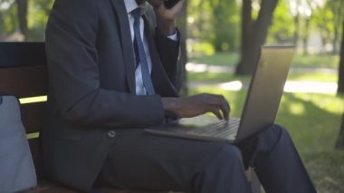 Güneş ışığında dizüstü bilgisayarda yazı yazan tanınmamış Afrikalı Amerikalı adam yaz parkında bankta oturuyormuş gibi. Çevrimiçi olarak çalışan meşgul genç bir iş adamı. Yaşam tarzı, iş, girişimcilik..