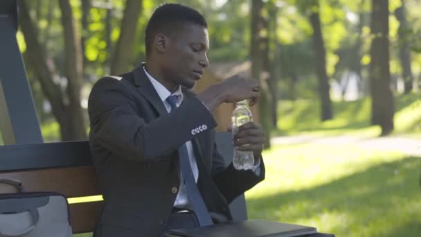 Cámara se acerca al hombre afroamericano feliz beber agua fría fresca al aire libre. Retrato de hombre de negocios confiado en traje descansando a la luz del sol en el parque de verano. Ocio, descanso, estilo de vida . — Vídeos de Stock