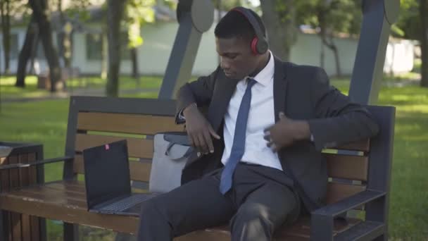 Joven hombre afroamericano guapo en traje poniéndose auriculares y escuchando música en el soleado parque de verano. Retrato de empresario serio disfrutando de hobby al aire libre. Estilo de vida, negocios . — Vídeo de stock