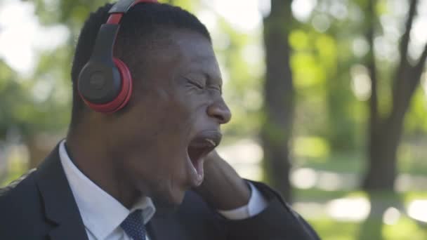 A vidám afro-amerikai férfi közelről élvezi a zenét a fejhallgatóban. Side view portré magabiztos üzletember elegáns öltönyben énekel hangosan a napos nyári parkban. Öröm, életmód, szabadidő. — Stock videók