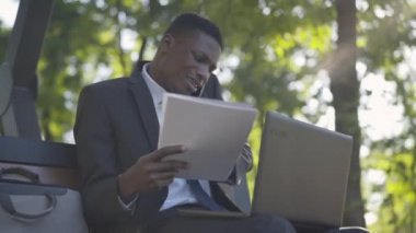 Tatmin olmamış Afro-Amerikan işadamının portresi güneşli bir parkta dizüstü bilgisayarla ve belgelerle oturuyor ve telefonda konuşuyor. Takım elbiseli stresli genç adam dışarıda çalışıyor. Aşırı çalışma, stres.