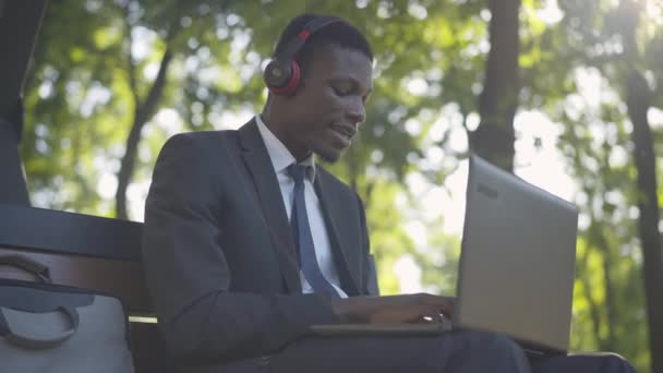 Alegre hombre afroamericano escuchando música y navegando por Internet. Retrato del alegre joven freelancer en traje de trabajo que trabaja en línea al aire libre en el soleado día de verano. Alegría, estilo de vida, libertad . — Vídeos de Stock