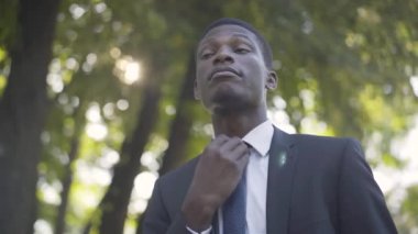 Genç Afrikalı Amerikalı adamın kravatını gevşetip iç çekişi. Güneşli parkta dinlenen yakışıklı başarılı iş adamının portresi. Yaşam tarzı, iş, rahatlama.