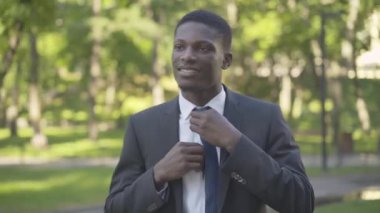 Takım elbiseli, kravatını düzelten ve kameraya gülümseyen kendine güvenen iş adamının orta pozu. Yaz parkında güneş ışığında poz veren genç bir Afrikalı Amerikalı. Yaşam tarzı, başarı, güven.