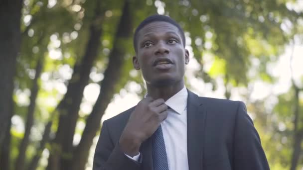 Close-up van de jonge knappe Afro-Amerikaanse man die stropdas aanpast in het zomerpark en vertrekt. Portret van een zelfverzekerde zakenman die loopt voor een ontmoeting. Zaken, levensstijl, vertrouwen, succes. — Stockvideo
