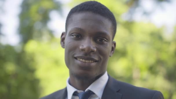 Close-up portrait of young African American man looking at camera and smiling. Positive businessman in formal suit posing outdoors in sunny summer park. Solitude, wealth, lifestyle. — Stock Video