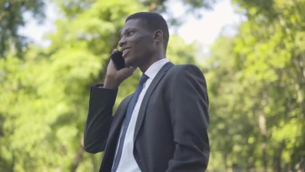 Portretul unui bărbat afro-american încrezător în costum vorbind la telefon în timp ce observă camera foto. Zâmbind tânăr om de afaceri stând în aer liber în parc în ziua însorită. Timp liber, fericire, stil de viață . — Videoclip de stoc