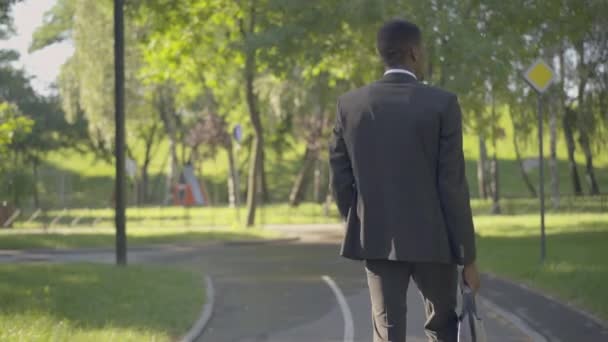 Rückenansicht eines erfolgreichen afroamerikanischen Geschäftsmannes, der an einem Sommertag im sonnigen Park spazieren geht. Die Kamera folgt dem selbstbewussten Mann im formellen Anzug, der in der Pause draußen spaziert. Wirtschaft, Lebensstil. — Stockvideo