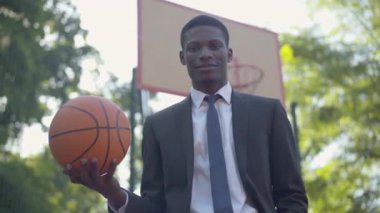 Basketbol sahasında topla poz veren zarif Afrikalı Amerikalı iş adamının portresi. Takım elbiseli, spora düşkün kendinden emin genç bir adamın orta pozu. Yaşam dengesi kavramı.