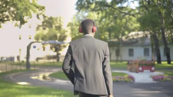 Die Kamera folgt dem fröhlichen, selbstbewussten Mann im Anzug, der mit Kopfhörern durch den Sommerpark läuft, sich dreht und singt. Porträt eines jungen Afroamerikaners, der an sonnigen Tagen im Freien Musik genießt. — Stockvideo