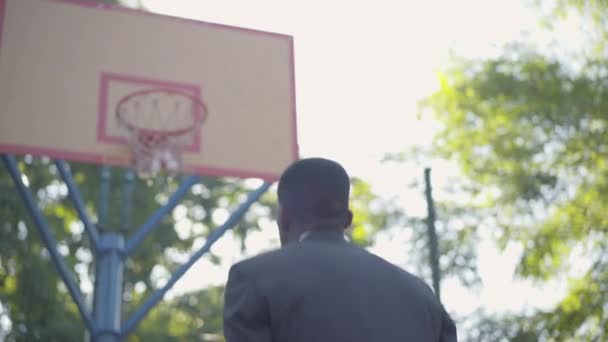 Vista posterior del hombre afroamericano en traje formal lanzando pelota en el aro y haciendo un gesto de victoria. Joven empresario confiado divirtiéndose como descansando al aire libre en un día soleado. Hobby, ocio . — Vídeos de Stock