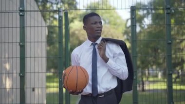 Takım elbiseli mutlu bir adamın portresi dışarıda basketbol sahasında topla yürüyüp gidiyor. Rahat Afrika kökenli Amerikalı işadamı güneşli bir yaz gününde dinleniyor. Hobi, spor.