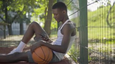 Afro-Amerikan basketbolcunun akıllı telefondan selfie çektiği bir yan görüntü portresi. Genç adam sahada oturuyor ve gülümsüyor. Hobi, spor, yaşam tarzı, atletizm.