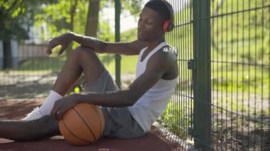 Pozitif Afrika kökenli Amerikalı sporcunun basketbol sahasında oturup kulaklıkla müzik dinlemesi. Güneşli yaz gününde tatilin tadını çıkaran yakışıklı genç adam. Spor konsepti.