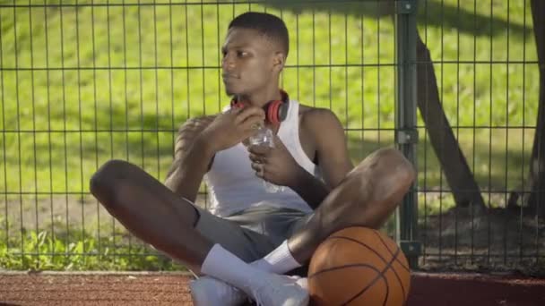 만족 스러운 농구 선수가 공을 들고 법정에 앉아 신선 한 냉수를 마시는 모습. 피곤 한 젊은 아프리카계 미국인 남자가 화창 한 날에 경기 후에 쉬는 모습이 보 입니다. 스포츠의 개념. — 비디오