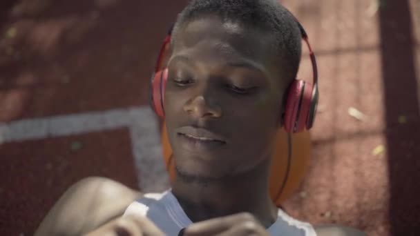 Cara de cerca del joven afroamericano acostado en la pelota de baloncesto y cantando. Retrato de deportista alegre en auriculares descansando al aire libre en un día soleado. Deporte, hobby, estilo de vida . — Vídeo de stock