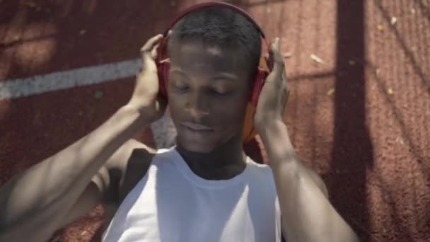 Portrait d'un sportif positif tenant un casque avec les mains et chantant. Vue du dessus du jeune homme afro-américain allongé sur un terrain de basket extérieur et profitant de la musique. — Video
