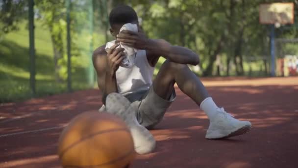 Agotado deportista afroamericano frotando la frente con una toalla blanca. Retrato de un joven cansado sentado en la cancha de baloncesto después de jugar al deporte. Deporte, resistencia, estilo de vida . — Vídeos de Stock