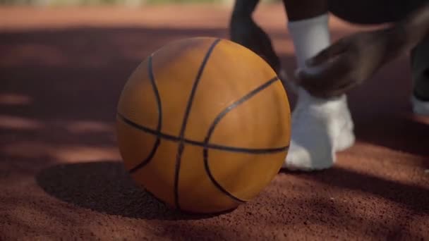 Неузнаваемый афроамериканский спортсмен завязывает шнурки на белых кроссовках, берёт мяч и уходит. Молодой человек играет в баскетбол. Спорт, здоровый образ жизни, атлетизм. — стоковое видео