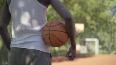Basketbol sahasında turuncu topla duran tanınmayan bir sporcunun arka görüntüsü. Spor giyimli genç Afrikalı Amerikalı adam güneşli bir günde açık havada spor oynuyor..