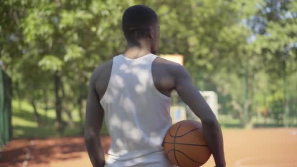 Rückenansicht eines afroamerikanischen Sportlers im weißen Hemd, der mit Ball auf einem sonnigen Basketballfeld steht, sich der Kamera zuwendet und lächelt. Junger selbstbewusster schöner Mann posiert an sonnigen Tagen im Freien. — Stockvideo
