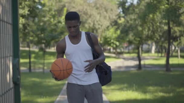 年轻自信的运动员在户外进入篮球场的画像。快乐的非裔美国人走路带球和微笑。生活方式、业余爱好、运动. — 图库视频影像