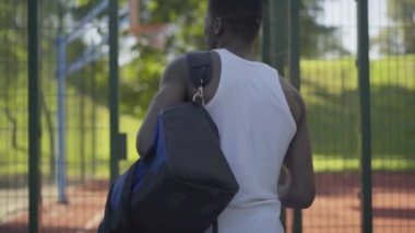 Spor çantalı ve toplu genç basketbolcunun sahaya çıkışının arka görüntüsü. Güneşli bir yaz gününde açık havada kendine güvenen Afrikalı Amerikalı bir adam rekabetçi bir oyun oynuyor..