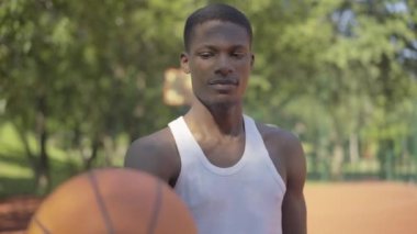 Afrika kökenli Amerikalı bir adamın kameraya topu uzatıp gülümsediği bir sahne. Basketbol sahasında poz veren kendinden emin genç bir sporcunun portresi. Eğlence, spor, yaşam tarzı.