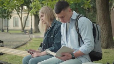 Kampüs avlusunda bankta oturup müzik eşliğinde dans eden rahat beyaz bir kız olarak kitap okuyan odaklanmış genç erkek öğrenci portresi. Üniversite yaşam tarzı, eğitim.