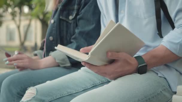 Närbild av oigenkännlig ung man sitter med bok utomhus och ung flicka som håller smartphone i bakgrunden. Två universitetsstudenter som studerar på skolgården. Utbildningsidé. — Stockvideo