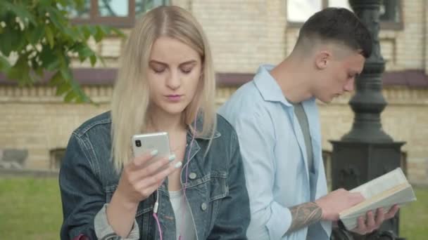 Retrato de una guapa rubia escuchando música en auriculares con un chico concentrado y borroso leyendo un libro al fondo. Pareja joven de estudiantes sentados al aire libre en el patio del campus universitario . — Vídeo de stock